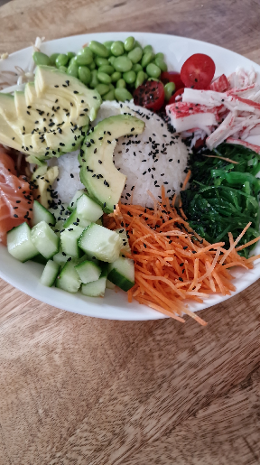 Een bord met Poké bowl, gevuld met Avocado, komkommer, wortel., adame boontjes, zalm, tonijn, surimi, tomaatjes en sushi rijst.
